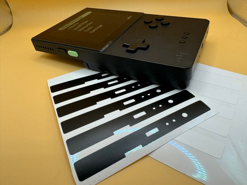 May include: A black handheld gaming console with a green button and a screen displaying the text "Library, Cart, Memories, Tools, Settings". The console is accompanied by a sheet of black and white stickers.