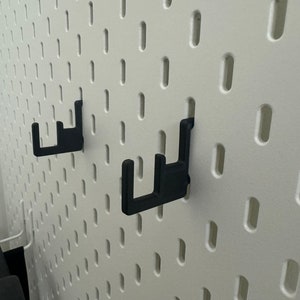 May include: Two black plastic hooks mounted on a white pegboard wall.