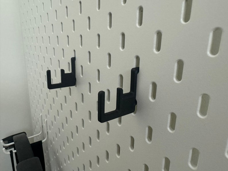 May include: Two black plastic hooks mounted on a white pegboard wall.