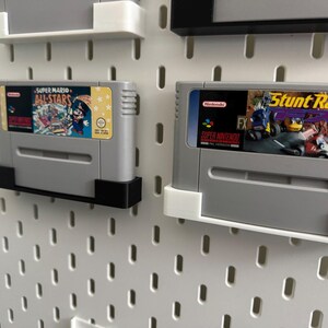 May include: Two grey Super Nintendo game cartridges, one labelled "Super Mario All-Stars" and the other "Stunt Race FX", mounted on a white pegboard. The cartridges are held by black and white brackets.