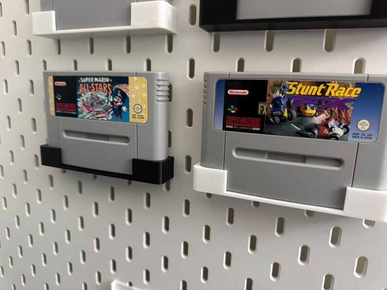May include: Two grey Super Nintendo game cartridges, one labelled "Super Mario All-Stars" and the other "Stunt Race FX", mounted on a white pegboard. The cartridges are held by black and white brackets.