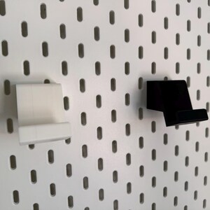 Op de afbeelding: Twee telefoonhouders voor pegboards, &eacute;&eacute;n witte en &eacute;&eacute;n zwarte, worden getoond tegen een witte pegboard achtergrond. De houders zijn ontworpen om een telefoon stevig vast te houden en zijn gemaakt van een stevig materiaal.