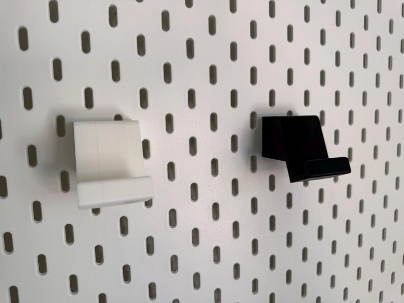 Op de afbeelding: Twee telefoonhouders voor pegboards, &eacute;&eacute;n witte en &eacute;&eacute;n zwarte, worden getoond tegen een witte pegboard achtergrond. De houders zijn ontworpen om een telefoon stevig vast te houden en zijn gemaakt van een stevig materiaal.