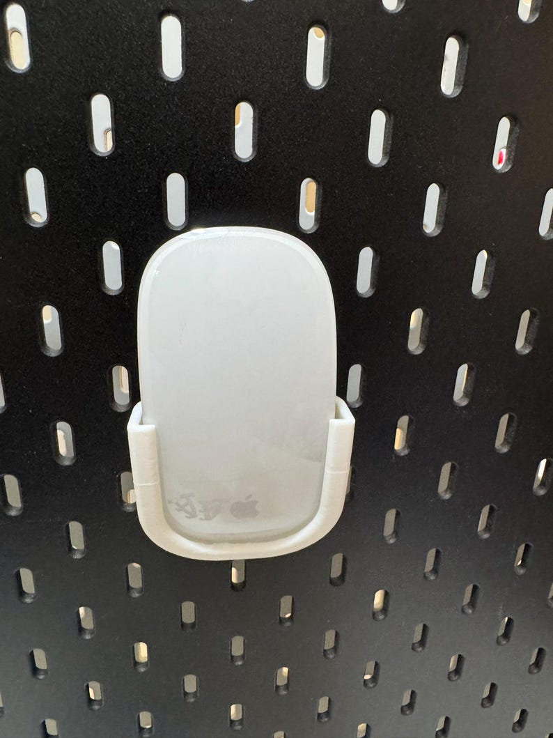 K&ouml;nnte beinhalten: Eine wei&szlig;e Computermaus wird in einer wei&szlig;en Kunststoffhalterung gehalten, die an einer schwarzen Lochwand befestigt ist. Die Maus hat ein glattes, abgerundetes Design und das Apple-Logo ist sichtbar. Die Lochwand hat gleichm&auml;&szlig;ig verteilte, ovale L&ouml;cher.