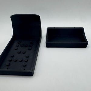 May include: Two black 3D-printed remote control holders. One is a rectangular tray with a curved back and a base with several raised circular features. The other is a smaller, rectangular tray with a curved front.