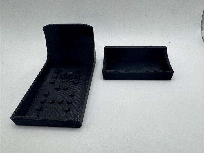 May include: Two black 3D-printed remote control holders. One is a rectangular tray with a curved back and a base with several raised circular features. The other is a smaller, rectangular tray with a curved front.