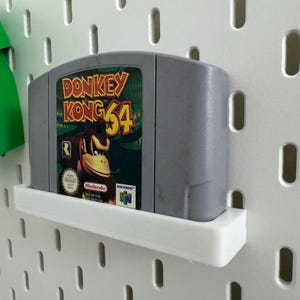 May include: A gray Nintendo 64 video game cartridge for Donkey Kong 64, mounted on a white shelf. The game's label features Donkey Kong and the game title in yellow and red. The shelf is attached to a white pegboard.