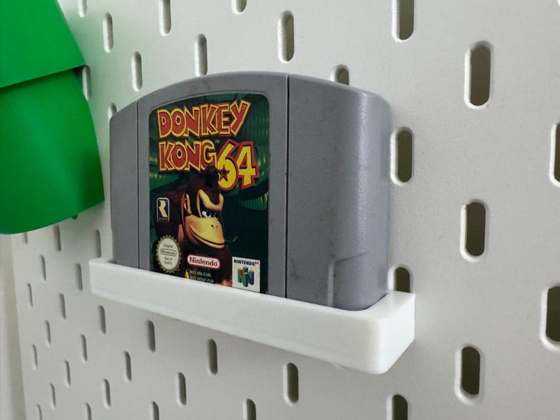 May include: A gray Nintendo 64 video game cartridge for Donkey Kong 64, mounted on a white shelf. The game's label features Donkey Kong and the game title in yellow and red. The shelf is attached to a white pegboard.