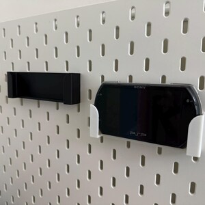 Pu&ograve; includere: Una pannello forato bianco con due supporti attaccati. Un supporto &egrave; nero e rettangolare. L'altro &egrave; bianco e contiene una console Sony PSP nera. La PSP &egrave; visibile con il testo "SONY" e "PSP" sullo schermo.