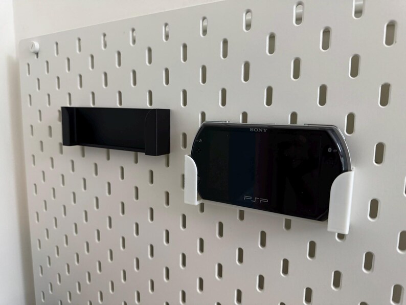 Pu&ograve; includere: Una pannello forato bianco con due supporti attaccati. Un supporto &egrave; nero e rettangolare. L'altro &egrave; bianco e contiene una console Sony PSP nera. La PSP &egrave; visibile con il testo "SONY" e "PSP" sullo schermo.