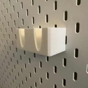 Puede incluir: Un organizador blanco rectangular para panel perforado con dos compartimentos curvos. El organizador est&aacute; montado en un panel perforado gris con agujeros ovalados espaciados. Dise&ntilde;ado para guardar objetos peque&ntilde;os.