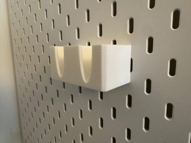 Puede incluir: Un organizador blanco rectangular para panel perforado con dos compartimentos curvos. El organizador est&aacute; montado en un panel perforado gris con agujeros ovalados espaciados. Dise&ntilde;ado para guardar objetos peque&ntilde;os.