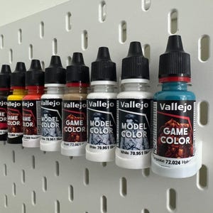 以下が含まれることがあります： 青、黄、赤、白、ターコイズなど、さまざまな色のVallejo Model ColorとGame Colorのペイントボトルを集めたものです。ボトルは白いペグボードに展示されており、黒いキャップとラベルが付いています。