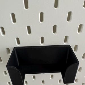Op de afbeelding: Zwarte plastic wandhouder voor een telefoon of ander klein apparaat. De houder is gemonteerd op een wit prikbord met gelijkmatig verdeelde gaten.