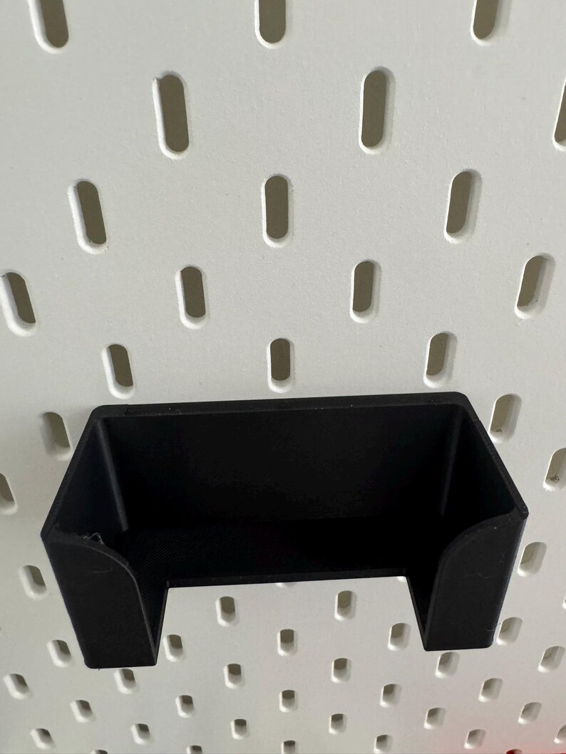Op de afbeelding: Zwarte plastic wandhouder voor een telefoon of ander klein apparaat. De houder is gemonteerd op een wit prikbord met gelijkmatig verdeelde gaten.