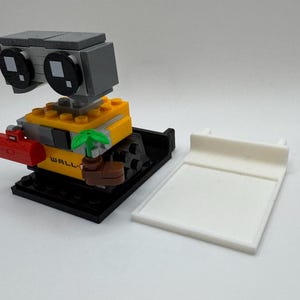 May include: A LEGO&reg; minifigure of WALL-E, a robot with a yellow body, black legs, and a gray head. The robot is holding a small plant and a red toolbox. The minifigure is standing on a black base with the word "WALL-E" printed on it.