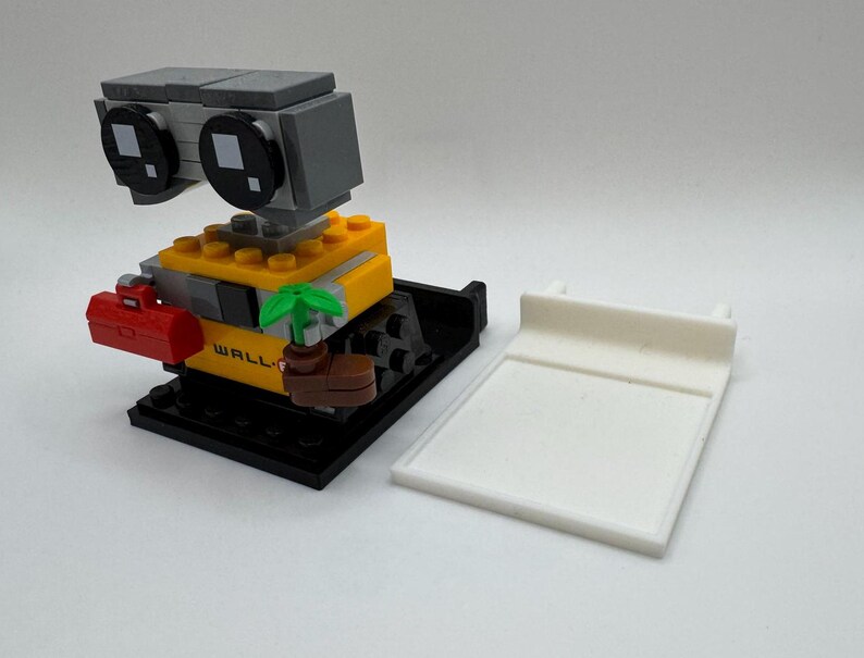 May include: A LEGO&reg; minifigure of WALL-E, a robot with a yellow body, black legs, and a gray head. The robot is holding a small plant and a red toolbox. The minifigure is standing on a black base with the word "WALL-E" printed on it.