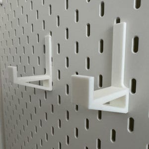 Op de afbeelding: Twee witte, 3D-geprinte pegboard haken zijn gemonteerd op een wit pegboard. De haken zijn L-vormig met een platte basis en een verticale steun. Het pegboard heeft gelijkmatig verdeelde gaten.