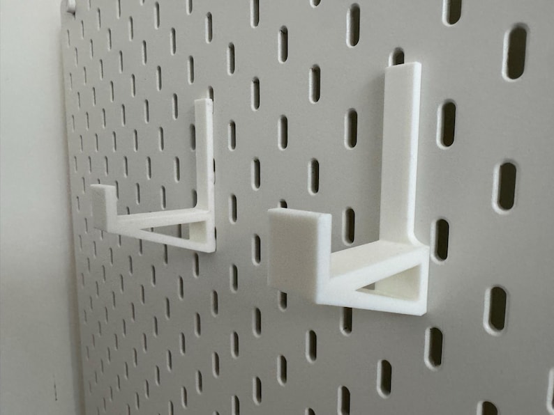Op de afbeelding: Twee witte, 3D-geprinte pegboard haken zijn gemonteerd op een wit pegboard. De haken zijn L-vormig met een platte basis en een verticale steun. Het pegboard heeft gelijkmatig verdeelde gaten.