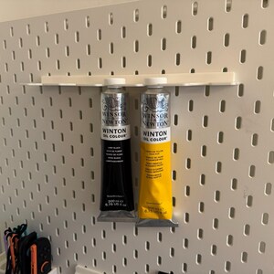 May include: Two tubes of Winsor & Newton Winton Oil Colour paint, one black and one yellow, displayed on a white pegboard. The black tube is labeled "Lamp Black" and the yellow tube is labeled "Yellow Ochre". Each tube contains 200 ml or 6.76 US fl oz.
