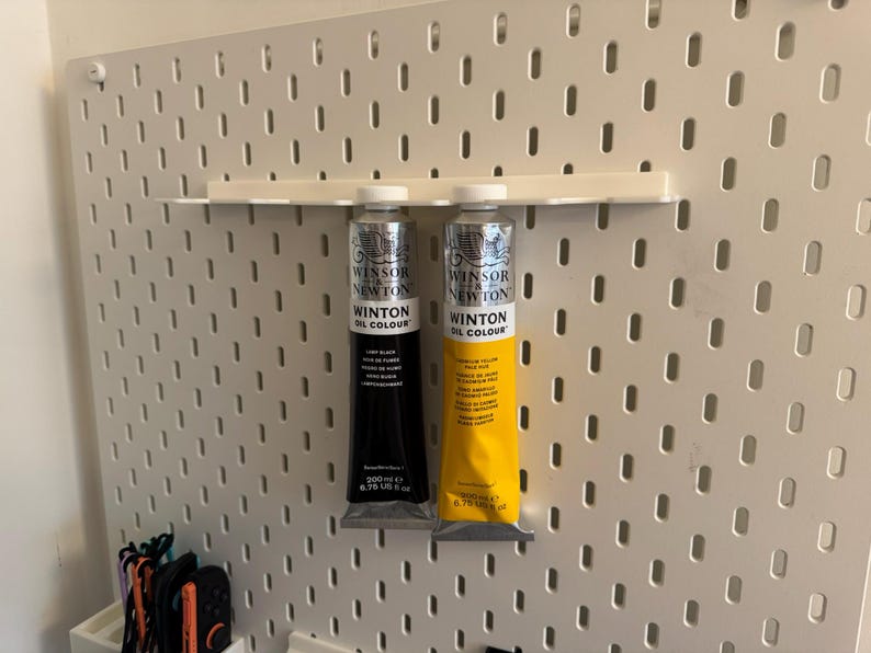 May include: Two tubes of Winsor & Newton Winton Oil Colour paint, one black and one yellow, displayed on a white pegboard. The black tube is labeled "Lamp Black" and the yellow tube is labeled "Yellow Ochre". Each tube contains 200 ml or 6.76 US fl oz.