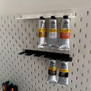 May include: A white pegboard displaying several tubes of Winsor & Newton Galeria acrylic paint. The silver tubes with black caps are held in place by black plastic holders. The paint tubes contain various colours.