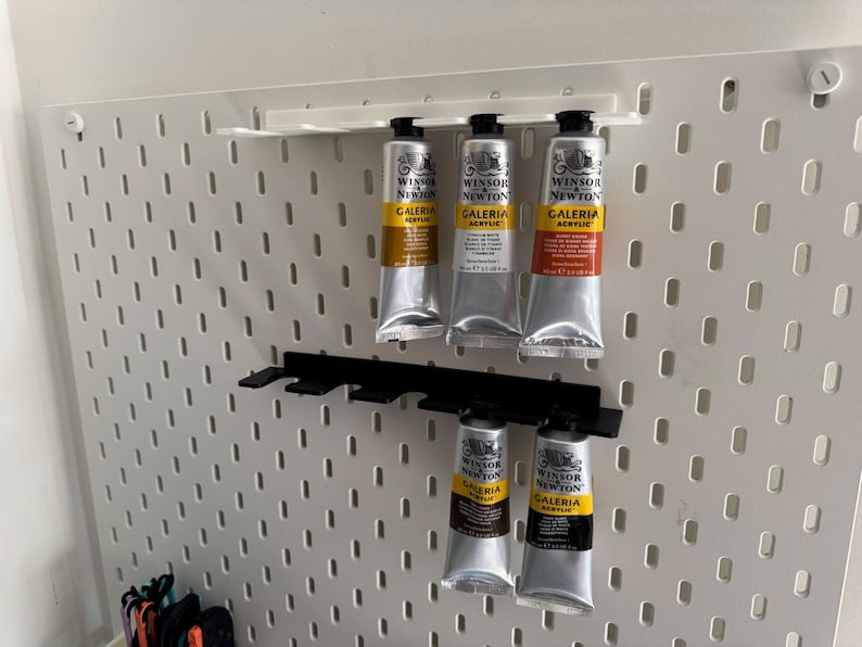 May include: A white pegboard displaying several tubes of Winsor & Newton Galeria acrylic paint. The silver tubes with black caps are held in place by black plastic holders. The paint tubes contain various colours.