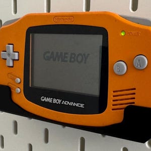 Op de afbeelding: Oranje Game Boy Advance draagbare videogame console met een zwarte wandbevestiging. De console heeft een grijs scherm met de tekst "GAME BOY" en "GAME BOY ADVANCE". De console heeft een groen stroomlampje.