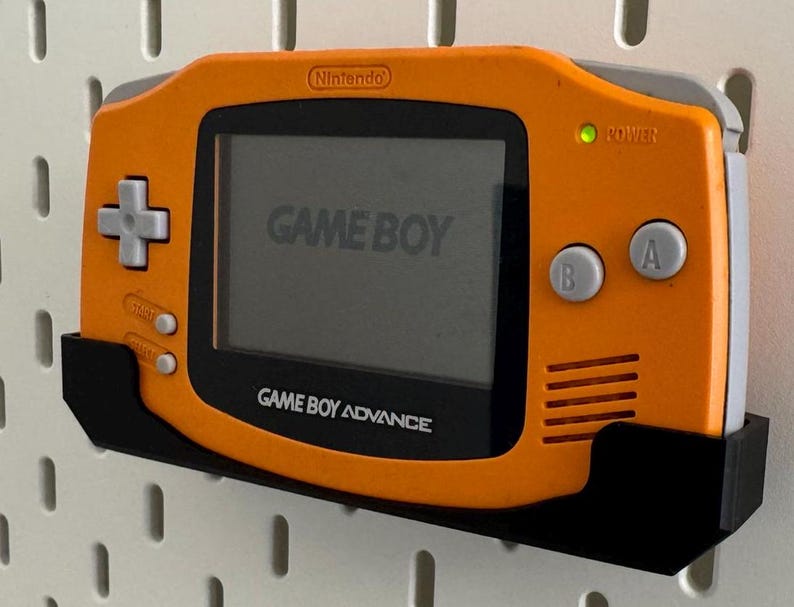 Op de afbeelding: Oranje Game Boy Advance draagbare videogame console met een zwarte wandbevestiging. De console heeft een grijs scherm met de tekst "GAME BOY" en "GAME BOY ADVANCE". De console heeft een groen stroomlampje.