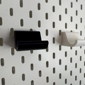 K&ouml;nnte beinhalten: Zwei Wandhalterungen f&uuml;r Telefone, eine schwarze und eine wei&szlig;e, werden vor einem wei&szlig;en Lochwandhintergrund gezeigt. Die Halterungen haben ein gebogenes Design, um ein Telefon oder ein &auml;hnliches Ger&auml;t aufzunehmen.