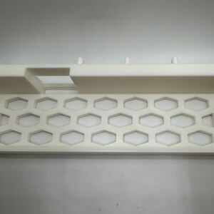 Puede incluir: Bandeja de pl&aacute;stico blanco con dise&ntilde;o de panal y borde elevado. La bandeja rectangular tiene una secci&oacute;n recortada y peque&ntilde;as clavijas a lo largo del borde superior. Probablemente para organizar art&iacute;culos.