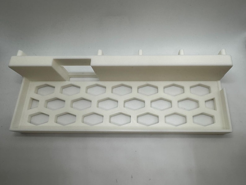 Puede incluir: Bandeja de pl&aacute;stico blanco con dise&ntilde;o de panal y borde elevado. La bandeja rectangular tiene una secci&oacute;n recortada y peque&ntilde;as clavijas a lo largo del borde superior. Probablemente para organizar art&iacute;culos.