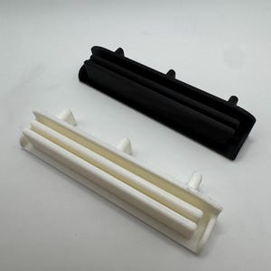 Pode incluir: Duas pe&ccedil;as de pl&aacute;stico impressas em 3D, uma preta e outra branca, s&atilde;o exibidas sobre um fundo branco. Cada pe&ccedil;a tem uma forma retangular com tr&ecirc;s cristas paralelas e pequenas protuber&acirc;ncias. As pe&ccedil;as parecem ser componentes de um produto maior.