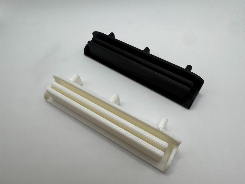 Pode incluir: Duas pe&ccedil;as de pl&aacute;stico impressas em 3D, uma preta e outra branca, s&atilde;o exibidas sobre um fundo branco. Cada pe&ccedil;a tem uma forma retangular com tr&ecirc;s cristas paralelas e pequenas protuber&acirc;ncias. As pe&ccedil;as parecem ser componentes de um produto maior.