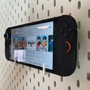 May include: Black Nintendo Switch console with a screen displaying game icons, mounted on a white pegboard. The console has black buttons and orange and blue accents. The screen shows game titles like Monopoly and Dragon Quest.