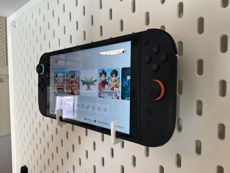 May include: Black Nintendo Switch console with a screen displaying game icons, mounted on a white pegboard. The console has black buttons and orange and blue accents. The screen shows game titles like Monopoly and Dragon Quest.