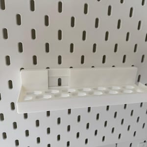 Puede incluir: Estante de pl&aacute;stico blanco con dise&ntilde;o rectangular y una serie de peque&ntilde;os agujeros redondos. Montado en un panel perforado blanco con agujeros ovalados espaciados. Dise&ntilde;ado para guardar objetos peque&ntilde;os.