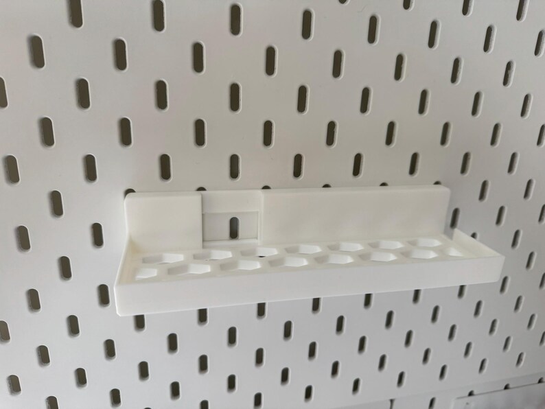 Puede incluir: Estante de pl&aacute;stico blanco con dise&ntilde;o rectangular y una serie de peque&ntilde;os agujeros redondos. Montado en un panel perforado blanco con agujeros ovalados espaciados. Dise&ntilde;ado para guardar objetos peque&ntilde;os.