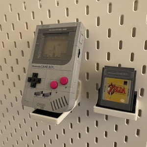May include: A gray Nintendo Game Boy handheld console with a game cartridge. The Game Boy has a black directional pad, pink buttons, and a screen displaying a game. The game cartridge features a gold and red label.
