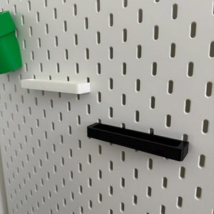 May include: A white pegboard with a series of evenly spaced, oval-shaped holes. Attached are three accessories: a green cup, a white shelf, and a black shelf. The accessories are designed to fit into the pegboard holes.