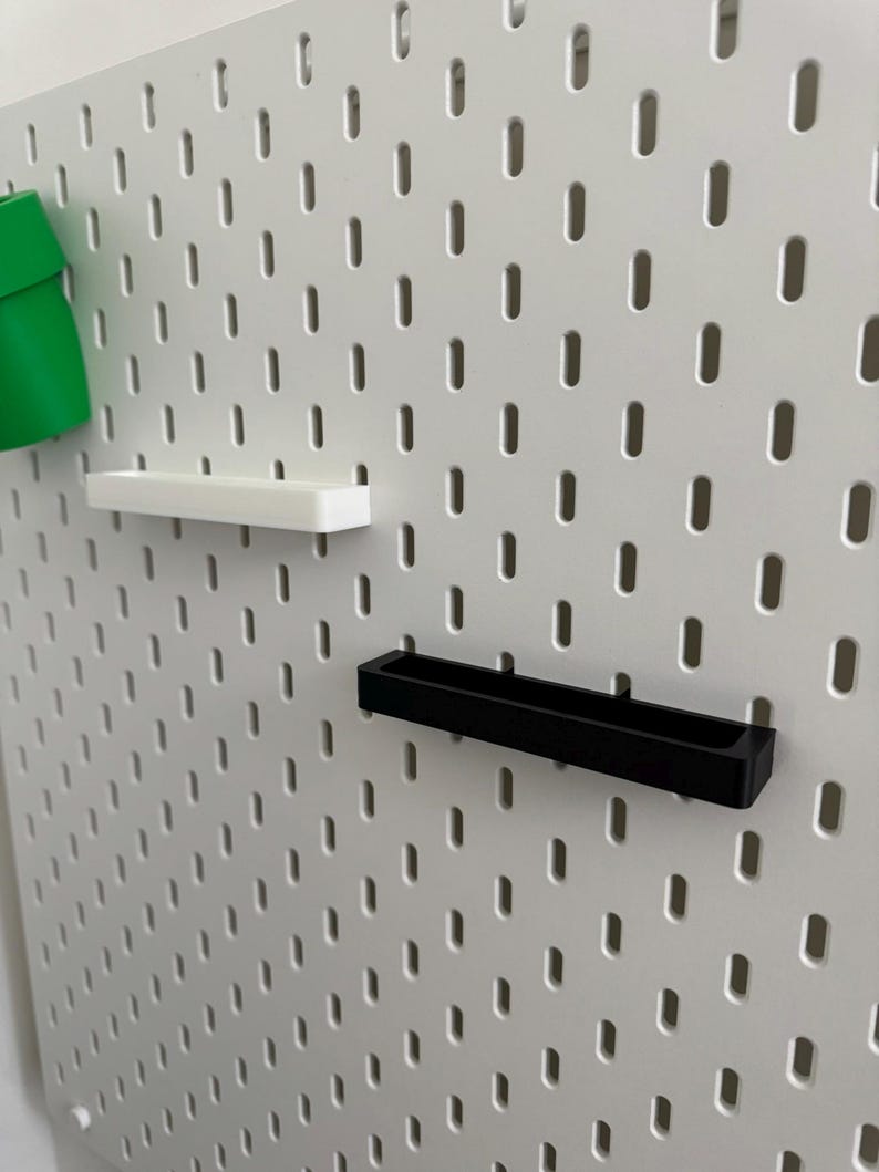 May include: A white pegboard with a series of evenly spaced, oval-shaped holes. Attached are three accessories: a green cup, a white shelf, and a black shelf. The accessories are designed to fit into the pegboard holes.