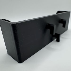 Op de afbeelding: Zwarte plastic wandbevestiging met twee kleine rechthoekige knoppen.