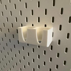 Puede incluir: Un organizador blanco impreso en 3D con dos compartimentos, montado en una pared perforada blanca. El organizador tiene aberturas curvas para facilitar el acceso. La pared perforada tiene una rejilla de agujeros ovalados espaciados.