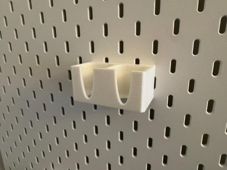 Puede incluir: Un organizador blanco impreso en 3D con dos compartimentos, montado en una pared perforada blanca. El organizador tiene aberturas curvas para facilitar el acceso. La pared perforada tiene una rejilla de agujeros ovalados espaciados.