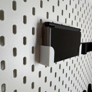 May include: A black handheld gaming console mounted on a white pegboard using white and black brackets. The pegboard has a pattern of evenly spaced, oval-shaped holes. The console is in a horizontal position.