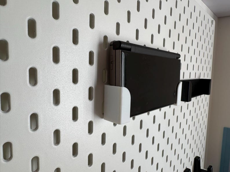 May include: A black handheld gaming console mounted on a white pegboard using white and black brackets. The pegboard has a pattern of evenly spaced, oval-shaped holes. The console is in a horizontal position.