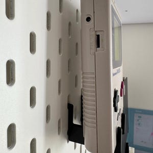 May include: A vintage off-white Nintendo Game Boy handheld console mounted on a white pegboard with black brackets. The Game Boy has a screen, buttons, and a cartridge slot. The pegboard has evenly spaced oval holes.