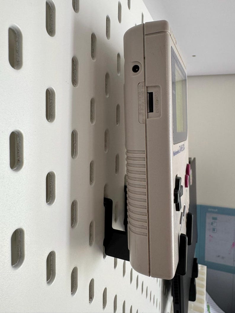 May include: A vintage off-white Nintendo Game Boy handheld console mounted on a white pegboard with black brackets. The Game Boy has a screen, buttons, and a cartridge slot. The pegboard has evenly spaced oval holes.