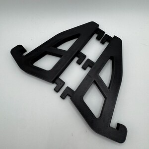 May include: A black, 3D-printed phone stand with an open, geometric design. The stand features two angled supports with a series of small, rectangular tabs for device placement. The stand is made of a textured material.