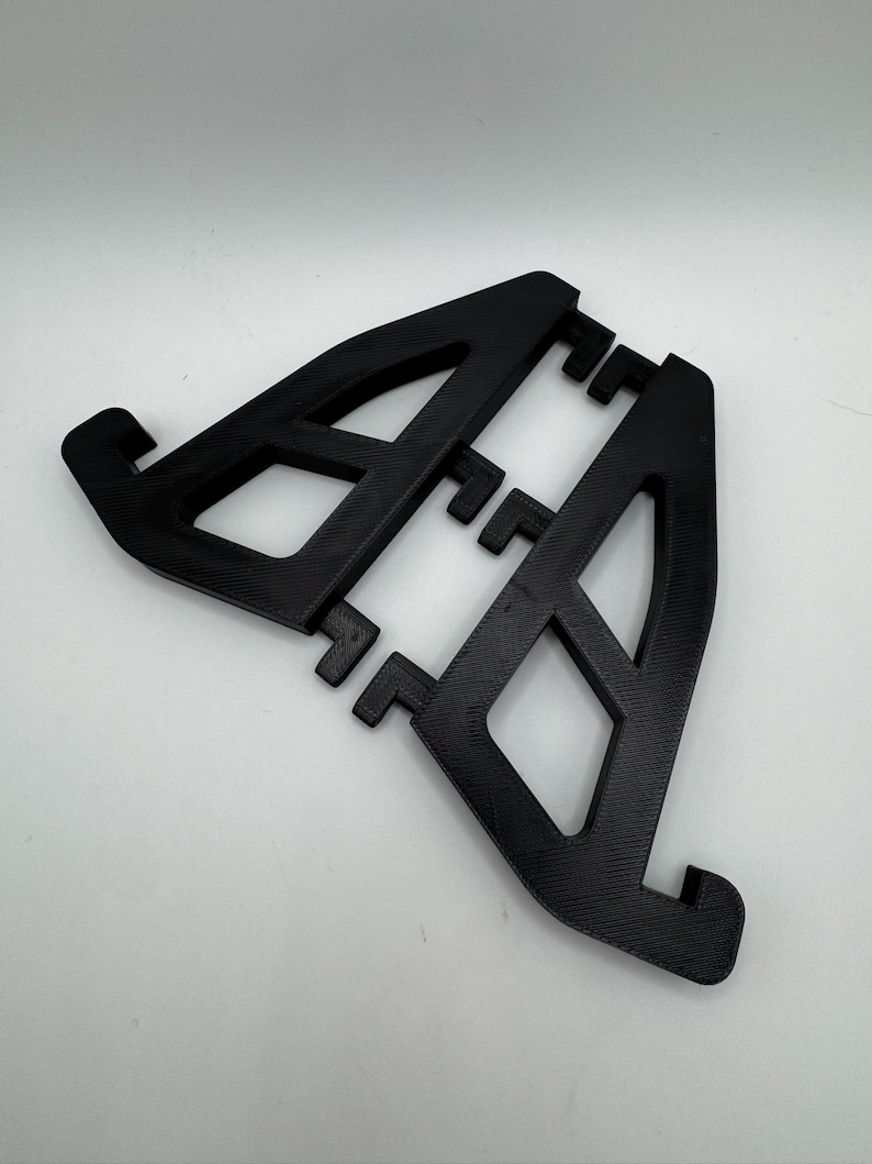 May include: A black, 3D-printed phone stand with an open, geometric design. The stand features two angled supports with a series of small, rectangular tabs for device placement. The stand is made of a textured material.